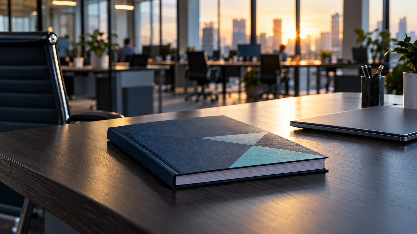 A dark blue bound volume lies on a dark wooden desk beside a closed laptop, with an open-plan office and a city skyline in low sun out of focus behind.