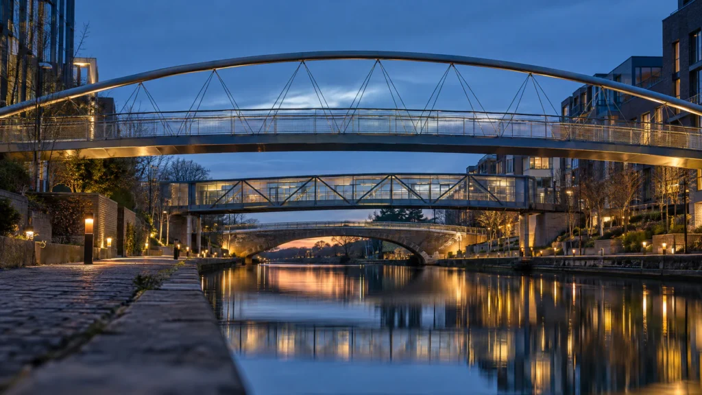 Three footbridges of different designs cross a calm city canal one behind another at dusk, seen from the towpath, with lights reflected in the water.