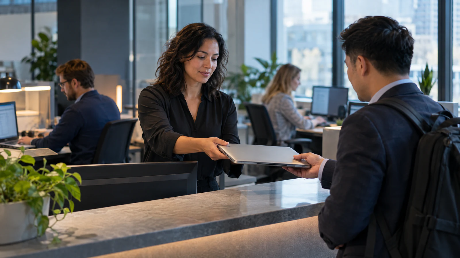 A woman behind an office service counter hands a closed laptop to a man with a backpack, while two colleagues work at desks behind her.